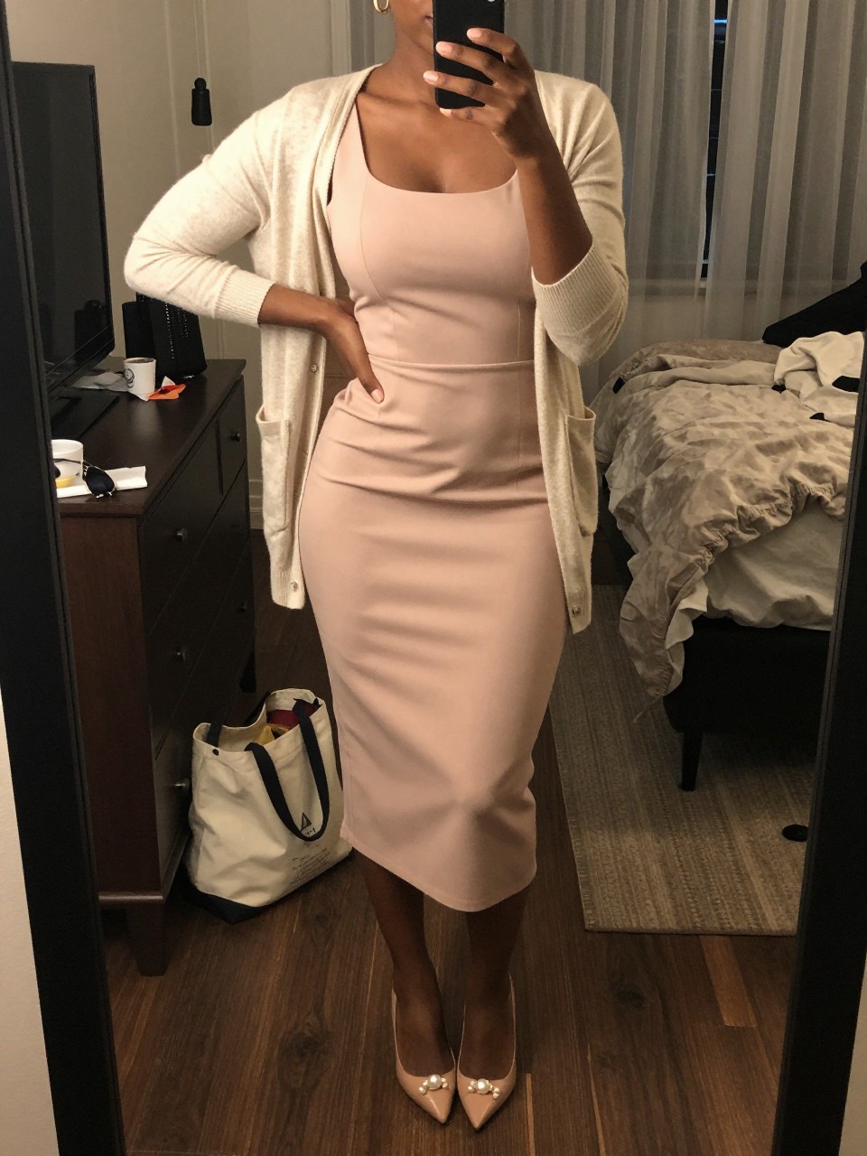 mirror selfie of a black woman wearing a blush crepe column midi dress with an ivory light cardigan, nude pointed-toe pumps, a pearl clutch, and small gold hoop earrings, holding a phone in front of a mirror, cropped at NECK so face doesn't show, with a natural casual selfie pose, in a city apartment, warm moody lighting, including small lived-in details like a tote bag on the floor, sunglasses on a dresser, or soft bedding in the background, latest iPhone photo quality.