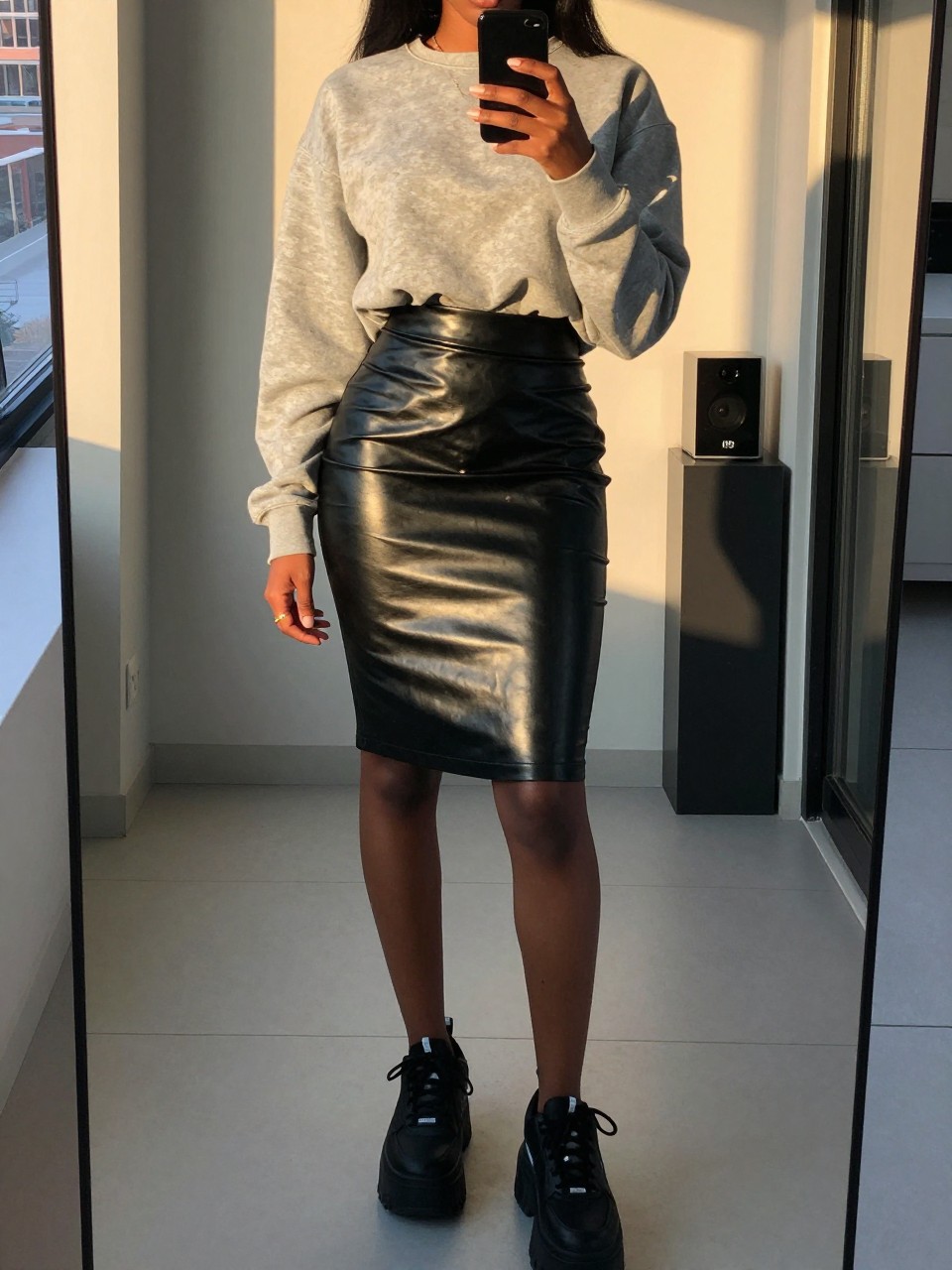 mirror selfie of a black woman wearing a gray sweatshirt half-tucked into a shiny black leather pencil skirt with chunky platform sneakers, standing with her phone in one hand, cropped at NECK so face doesn't show, in a city apartment, evening golden hour, iPhone photo quality.