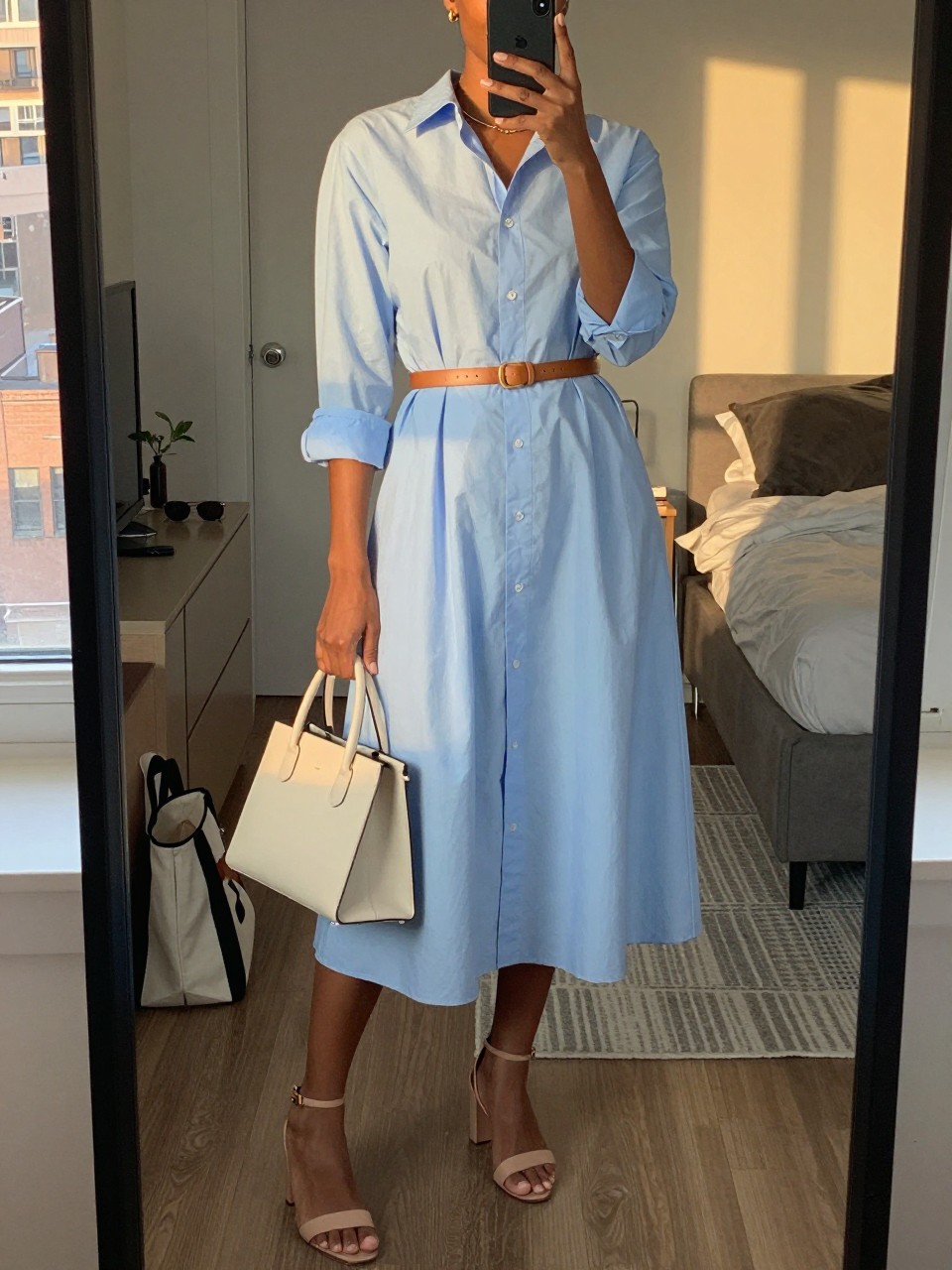 mirror selfie of a black woman wearing a pale blue poplin midi shirt dress with a tan leather waist belt, nude block-heel sandals, a cream structured handbag, and simple gold stud earrings, holding a phone in front of a mirror, cropped at NECK so face doesn't show, with a natural casual selfie pose, in a city apartment, evening golden hour, including small lived-in details like a tote bag on the floor, sunglasses on a dresser, or soft bedding in the background, latest iPhone photo quality.