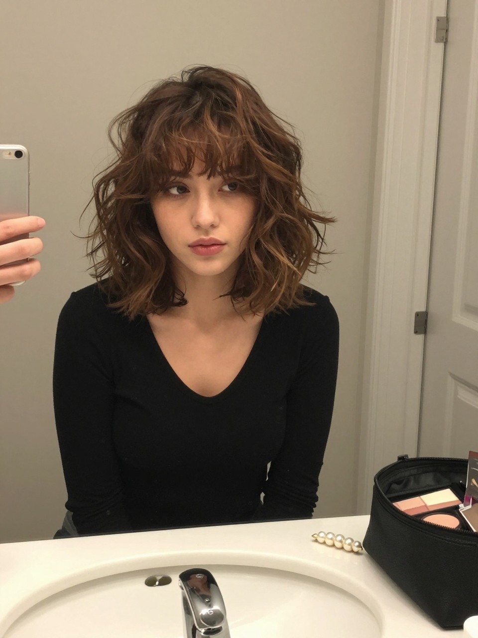 Photo of a woman wearing a short brown lob with chestnut highlights and soft brushed curls, candid mirror selfie, at a bathroom vanity mirror, soft indoor lighting, containing a small lived-in detail such as a pearl clip near the sink and a makeup bag left open nearby, latest iPhone photo quality.