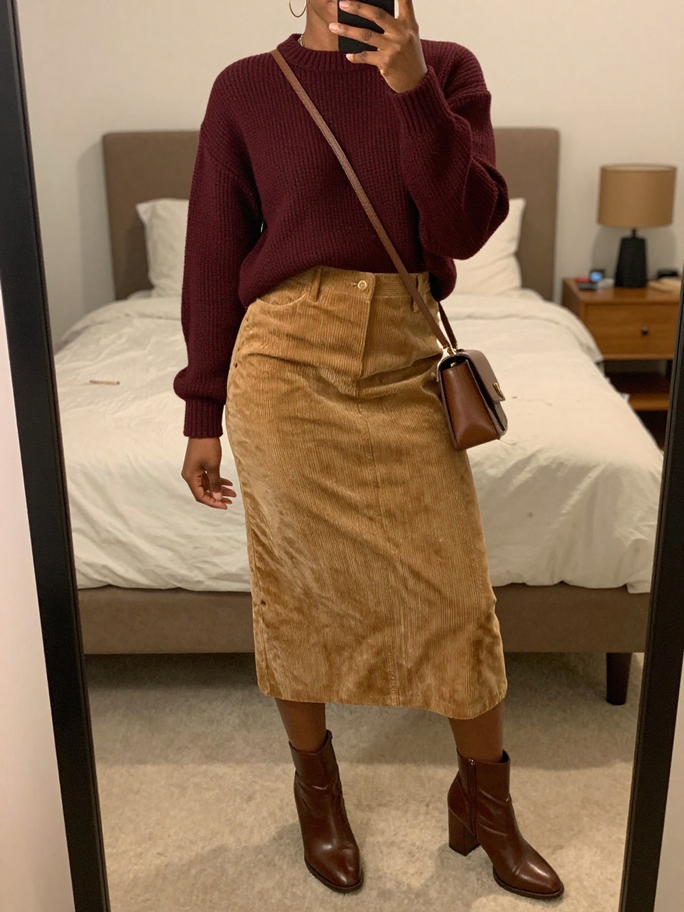 mirror selfie of a black woman wearing a deep burgundy fine-knit sweater tucked into a tan brown corduroy midi skirt with brown leather block-heel ankle boots and a matching brown crossbody bag, large gold hoops visible, standing in a confident pose, cropped at NECK so face doesn't show, in a feminine-themed bedroom, warm moody lighting, iPhone photo quality.