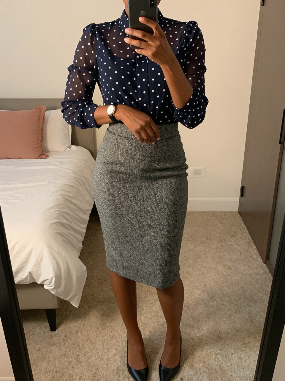 mirror selfie of a black woman wearing a sheer navy polka-dot blouse tucked into a gray herringbone pencil skirt with pointed flats, one hand holding her phone, the other adjusting her watch, cropped at NECK so face doesn't show, in a feminine-themed bedroom, evening golden hour, iPhone photo quality.