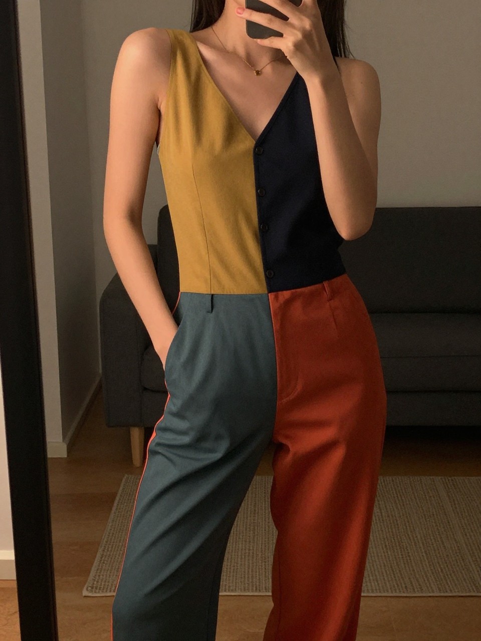 iPhone mirror selfie of a woman in a color-block jumpsuit, posing with hands in pockets, cropped at NECK so face doesn't show, in a city apartment, warm moody lighting, iPhone photo quality.