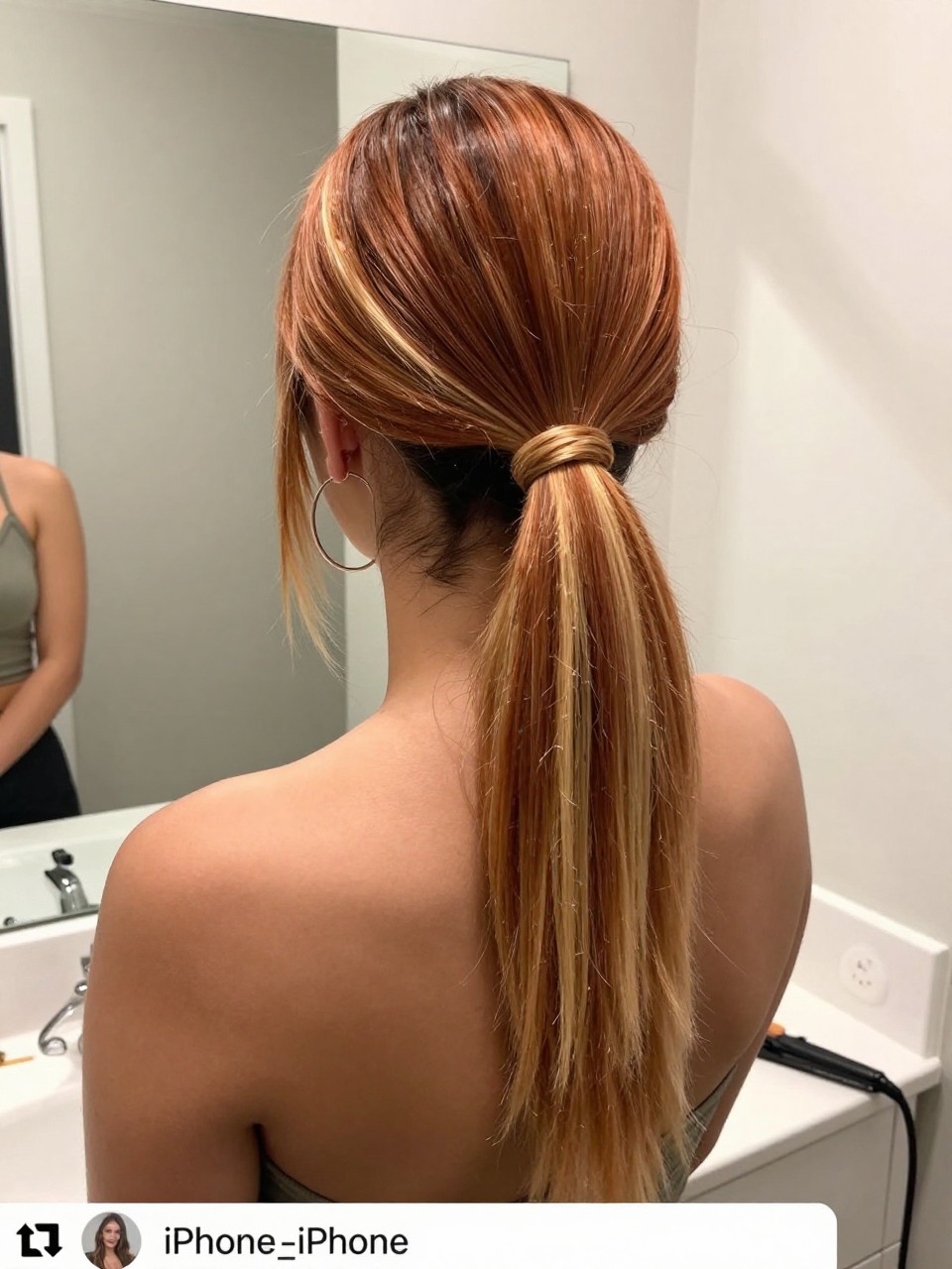 Photo of a mixed woman wearing a sleek mid ponytail with copper hair and blonde highlight panels through the tail, back-of-head view to highlight details, at a bathroom vanity mirror, soft indoor lighting, containing a small lived-in detail such as a flat iron on the counter and hoop earrings peeking through, latest iPhone photo quality.
