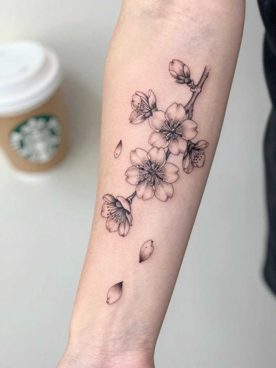 Image Prompt: "Close-up of a Japanese plum blossom tattoo with falling petals on the forearm, fine line black and grey ink, soft shading, delicate drifting petal details, realistic skin texture, coffee cup softly blurred nearby, feminine lifestyle touch kept minimal, tattoo-centered inspiration image, soft clean background (no watermarks on images)"