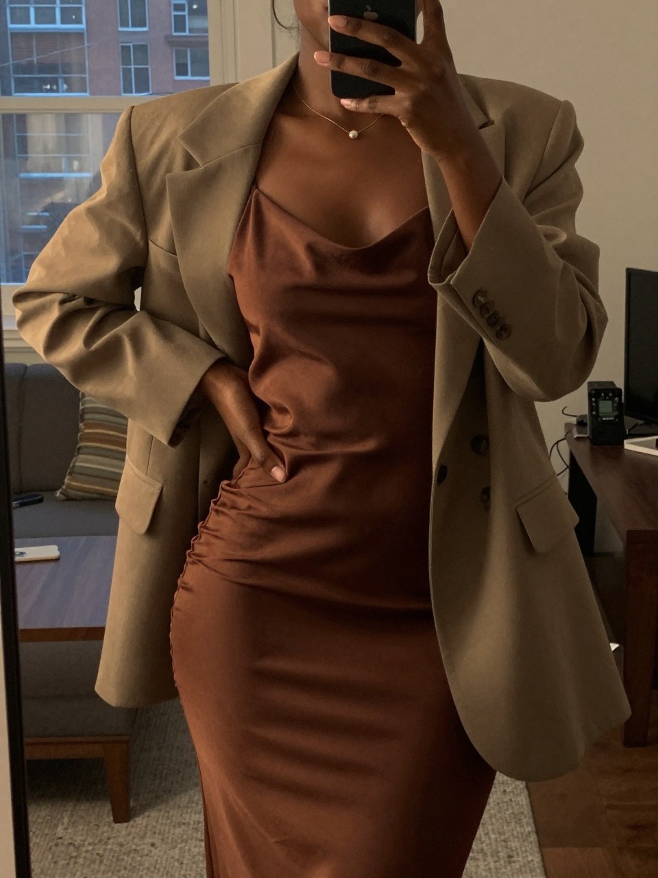 iPhone mirror selfie of a black woman in a slip dress with a blazer over her shoulders, hand on hip, cropped at NECK so face doesn't show, in a city apartment, warm moody lighting, iPhone photo quality.