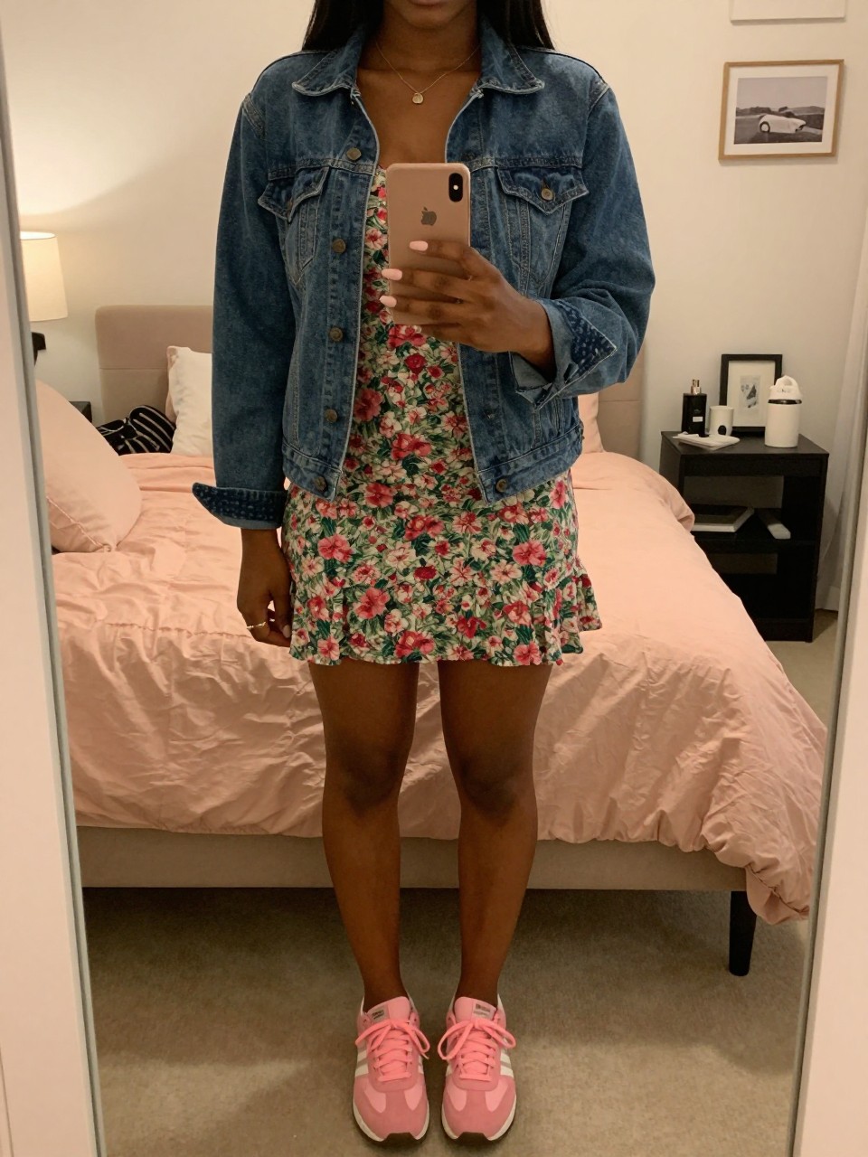 mirror selfie of a black woman in a floral mini dress and denim jacket with pink sneakers, holding phone at waist, cropped at NECK so face doesn't show, in a feminine-themed bedroom, warm moody lighting, iPhone photo quality.