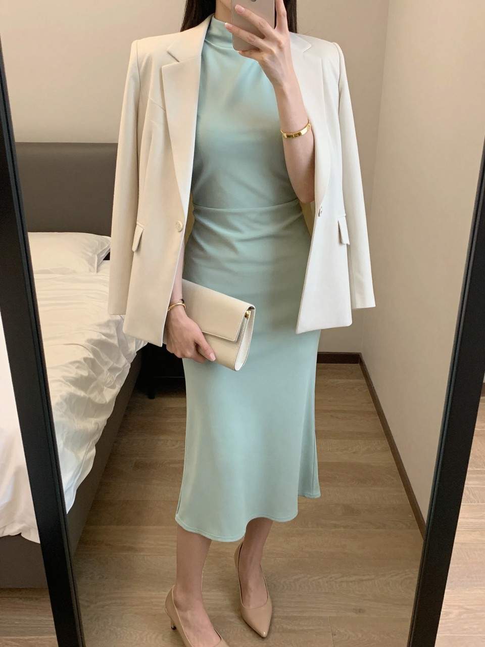 mirror selfie of a woman wearing a soft mint green high-neck midi dress with an ivory light blazer, nude slingback pumps, a clean gold cuff bracelet, and a cream structured clutch, holding a phone in front of a mirror, cropped at NECK so face doesn't show, with a natural casual selfie pose, in a feminine-themed bedroom, warm moody lighting, including small lived-in details latest iPhone photo quality.