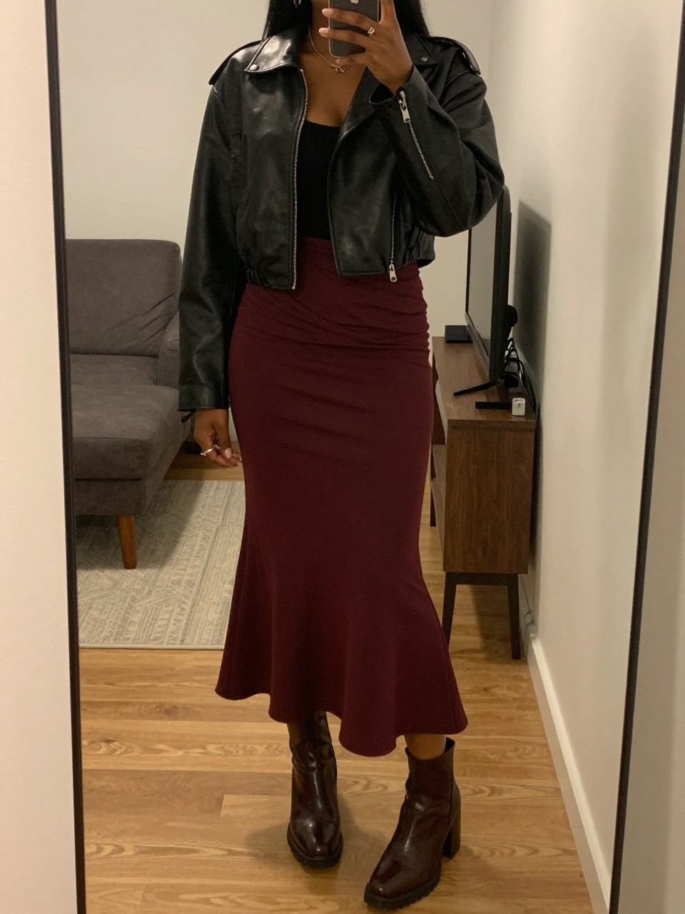 mirror selfie of a black woman in a cropped black leather jacket and a burgundy mermaid skirt with chunky ankle boots, one hand in her jacket pocket, cropped at NECK so face doesn't show, in a city apartment, warm moody lighting, iPhone photo quality.