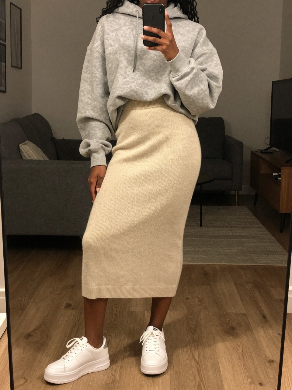 iPhone mirror selfie of a black woman wearing an oversized gray hoodie half-tucked into a soft cream knit midi skirt with chunky white platform sneakers, one hand holding her phone low by her thigh, cropped at NECK so face doesn't show, in a city apartment, warm moody lighting, iPhone photo quality.