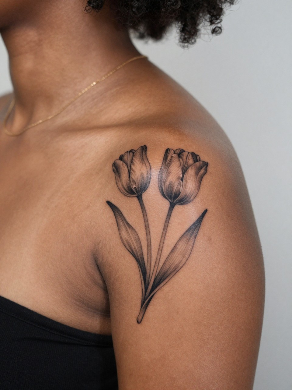 Image Prompt: "Close-up of a fine line tulip pair tattoo on the shoulder of a black feminine woman, black ink floral design with smooth petals, slender stems, subtle soft grey shading, realistic skin texture, delicate gold necklace chain faintly visible, feminine modern tattoo inspiration image, minimal clean background, tattoo is the clear main focus (no watermarks on images)"