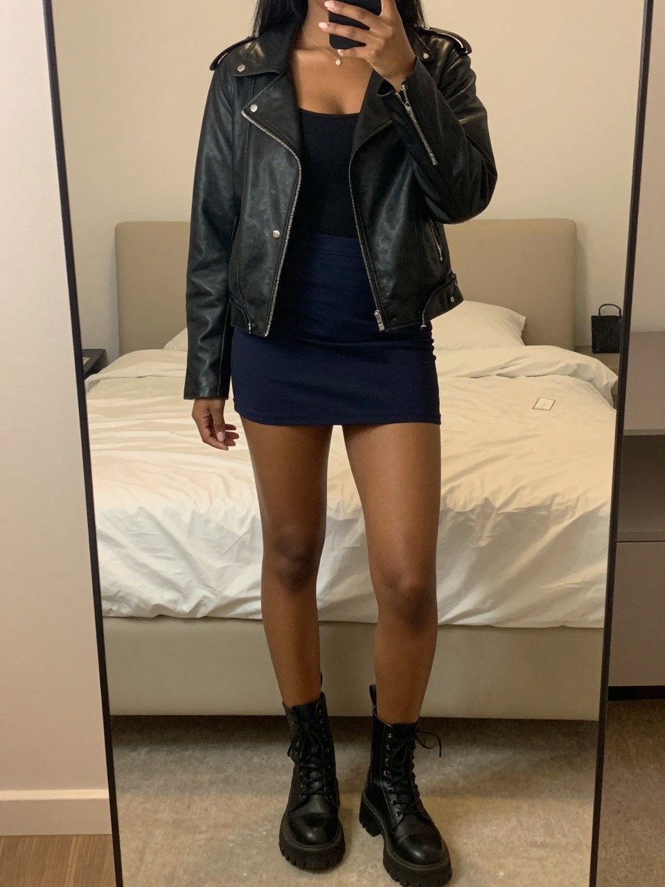 iPhone mirror selfie of a black woman wearing a black leather jacket over a black tank top with a navy mini skirt and chunky combat boots, one hand in her jacket pocket, cropped at NECK so face doesn't show, in a feminine-themed bedroom, warm moody lighting, iPhone photo quality.