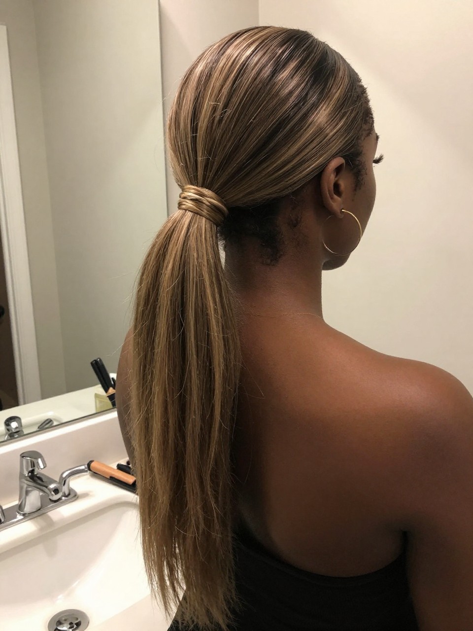 Photo of a black woman wearing a sleek mid ponytail with ash brown hair and ash blonde highlight panels through the tail, back-of-head view to highlight details, at a bathroom vanity mirror, soft indoor lighting, containing a small lived-in detail such as a flat iron on the counter and hoop earrings peeking through, latest iPhone photo quality.