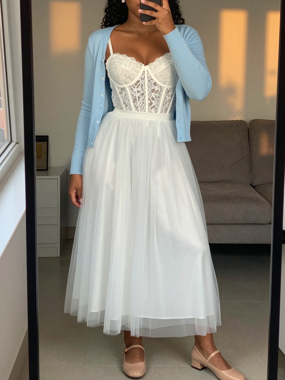 mirror selfie of a curvy black woman wearing a white lace corset top with a high-waisted white tulle maxi skirt, a baby blue cardigan draped over her shoulders, and nude Mary Jane flats, pearl earrings visible, standing in a soft romantic pose, cropped at NECK so face doesn't show, in a city apartment, evening golden hour, iPhone photo quality.