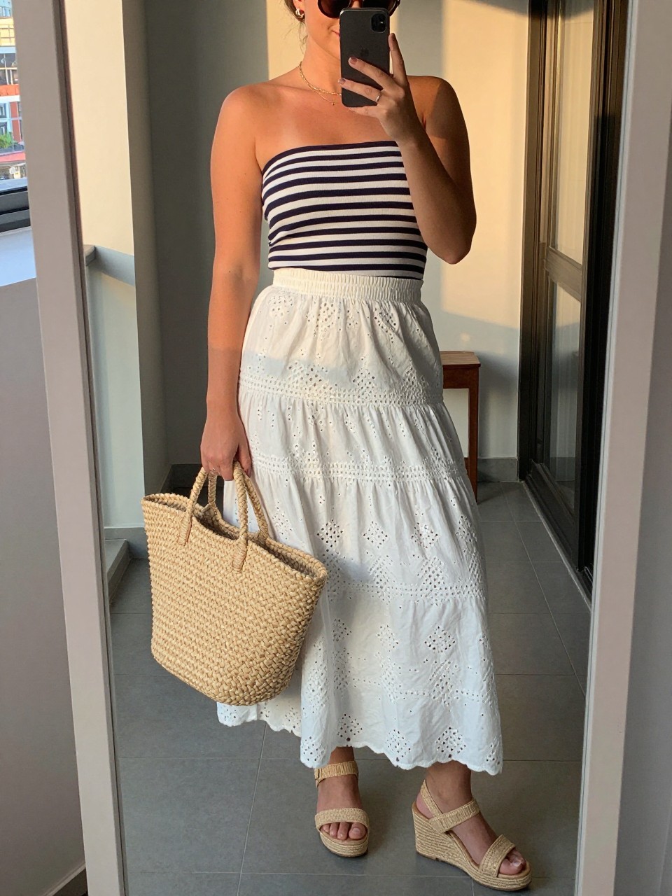 mirror selfie of a curvy woman wearing a navy and white striped boatneck top tucked into a white eyelet maxi skirt with raffia wedge sandals and a large straw tote bag, sunglasses perched on her head, standing in a relaxed summer pose, cropped at NECK so face doesn't show, in a city apartment, evening golden hour, iPhone photo quality.