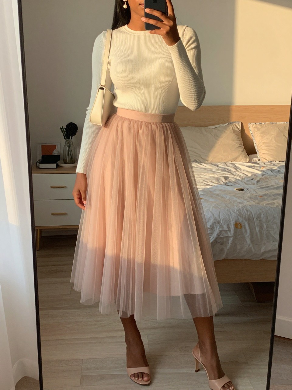 mirror selfie of a black woman wearing a blush tulle midi skirt with a cream ribbed fitted knit top, nude heeled mules, pearl drop earrings, and an ivory mini shoulder bag, holding a phone in front of a mirror, cropped at NECK so face doesn't show, with a natural casual selfie pose, in a feminine-themed bedroom, evening golden hour, including small lived-in details latest iPhone photo quality.