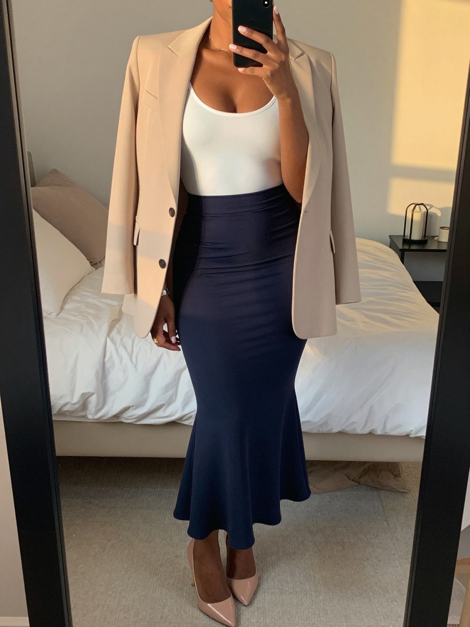 mirror selfie of a black woman in a white bodysuit and beige blazer with a navy mermaid skirt and nude pumps, standing with her phone in hand, cropped at NECK so face doesn't show, in a feminine-themed bedroom, evening golden hour, iPhone photo quality.