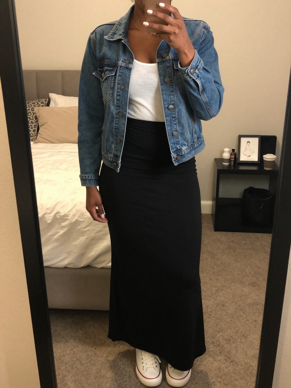 iPhone mirror selfie of a black woman in a black maxi skirt and denim jacket over a white tank with sneakers, standing with hand in jacket pocket, cropped at NECK so face doesn't show, in a feminine-themed bedroom, warm moody lighting, iPhone photo quality.