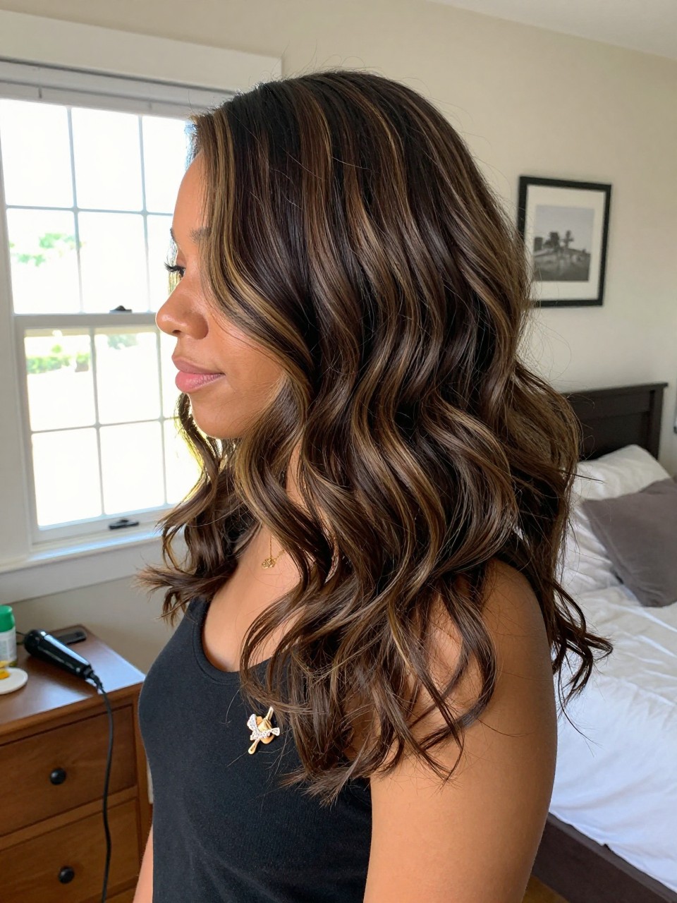 Photo of a biracial woman wearing soft brown waves with dark chestnut highlight streaks, side profile view, in a sun-drenched bedroom, natural window light, containing a small lived-in detail such as a decorative pin on one side and a curling iron resting on the nightstand, latest iPhone photo quality.