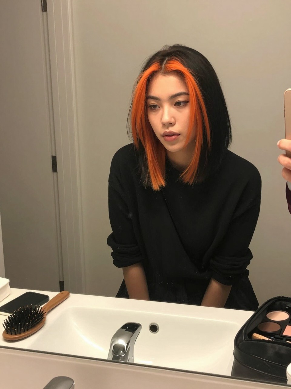 Photo of a woman wearing a sleek black lob with bright orange face-framing highlights, candid mirror selfie, at a bathroom vanity mirror, soft indoor lighting, containing a small lived-in detail such as a hairbrush on the vanity and a makeup bag nearby, latest iPhone photo quality.