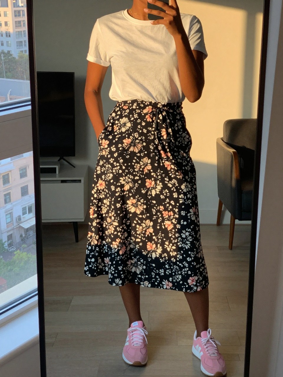 iPhone mirror selfie of a black woman in a floral midi skirt and white tee with pink sneakers, standing with hand in pocket, cropped at NECK so face doesn't show, in a city apartment, evening golden hour, iPhone photo quality.