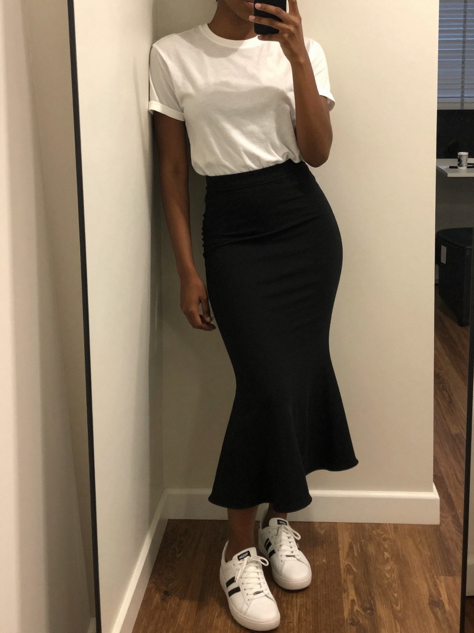 mirror selfie of a black woman in a white tee and a black cotton mermaid skirt with white sneakers, casually leaning against a wall, cropped at NECK so face doesn't show, in a city apartment, warm moody lighting, iPhone photo quality.