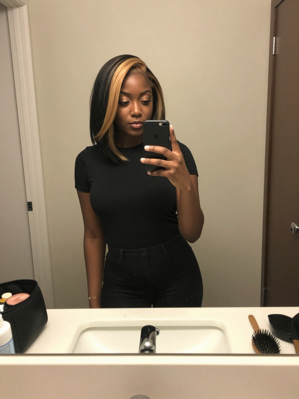 Photo of a black woman wearing a sleek black lob with blonde face-framing highlights, candid mirror selfie, at a bathroom vanity mirror, soft indoor lighting, containing a small lived-in detail such as a hairbrush on the vanity and a makeup bag nearby, latest iPhone photo quality.