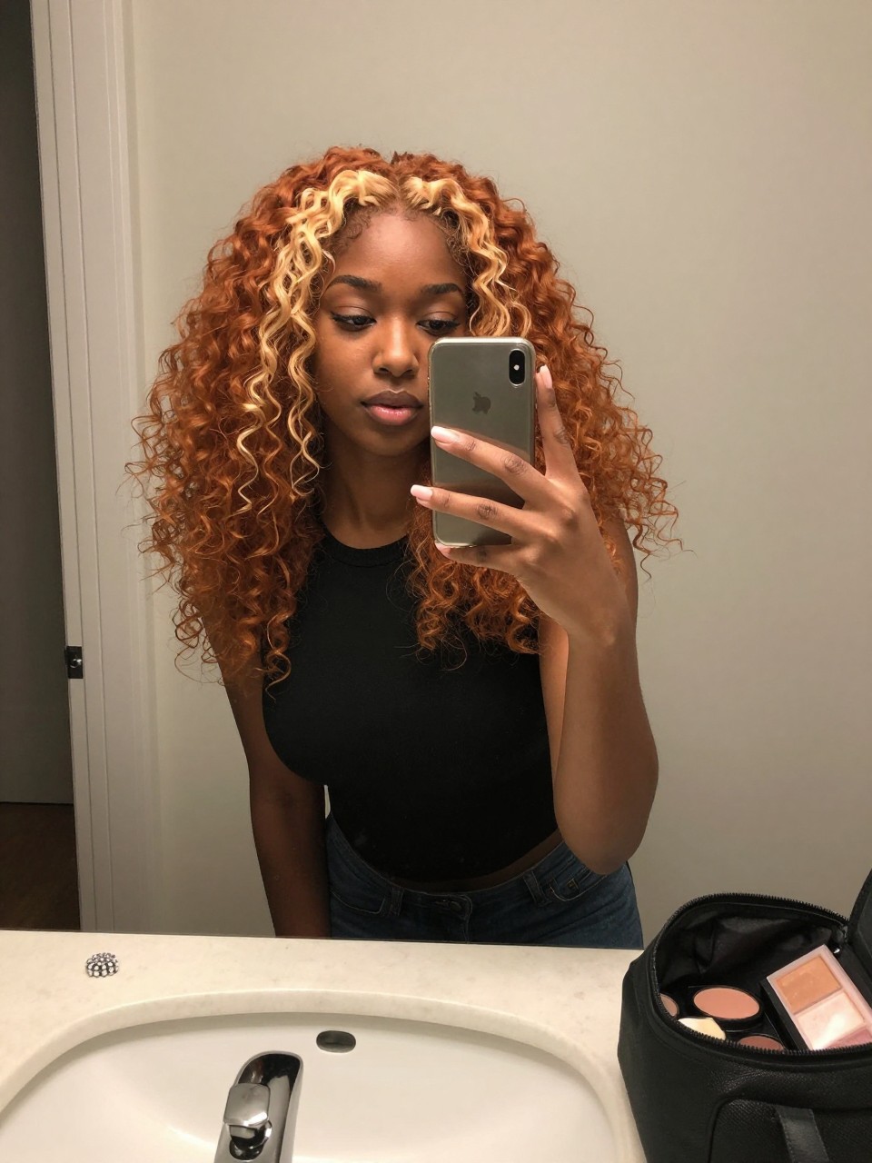 Photo of a black woman wearing soft curled ginger hair with creamy gold highlights throughout, candid mirror selfie, at a bathroom vanity mirror, soft indoor lighting, containing a small lived-in detail such as a rhinestone clip near the sink and a makeup bag left open nearby, latest iPhone photo quality.