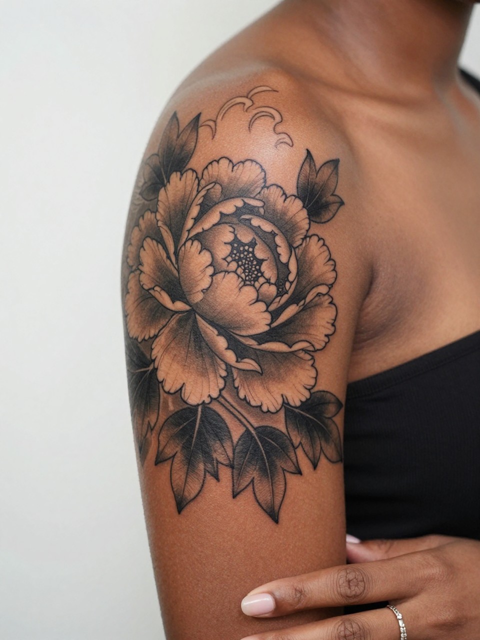 Image Prompt: "Close-up of a Japanese peony tattoo with subtle wave detail on the upper arm of a black woman, black and grey ink, layered petals, soft shading, traditional-inspired linework, realistic skin texture, delicate ring visible on hand near frame, feminine tattoo inspiration photography, minimal background, tattoo is the hero (no watermarks on images)"