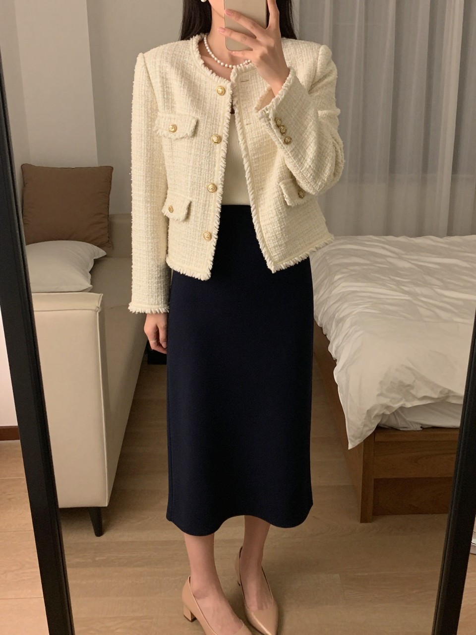 mirror selfie of a woman wearing a cream tweed jacket over a cream shell tucked into a navy midi skirt with pearl earrings and nude pumps, one hand resting on her jacket, cropped at NECK so face doesn't show, in a feminine-themed bedroom, warm moody lighting, iPhone photo quality.