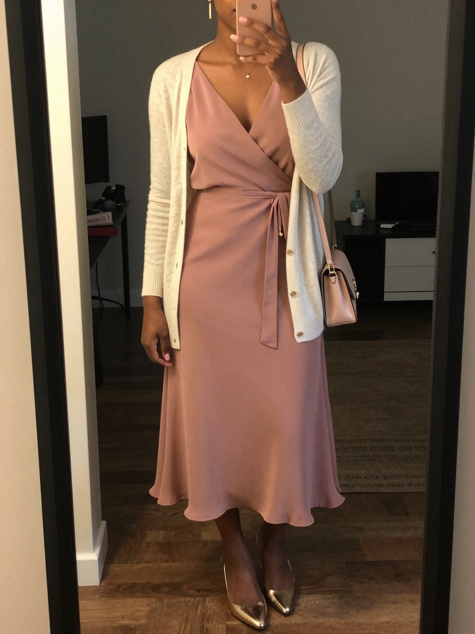 mirror selfie of a black woman wearing a dusty rose chiffon wrap midi dress with a cream light cardigan, metallic gold heels, simple gold drop earrings, and a blush small shoulder bag, holding a phone in front of a mirror, cropped at NECK so face doesn't show, with a natural casual selfie pose, in a city apartment, warm moody lighting, including small lived-in details, latest iPhone photo quality.