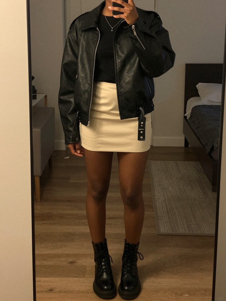 mirror selfie of a black woman wearing a black leather jacket over a band tee with a soft cream mini skirt and black combat boots, one hand in her jacket pocket, cropped at NECK so face doesn't show, in a city apartment, warm moody lighting, iPhone photo quality.