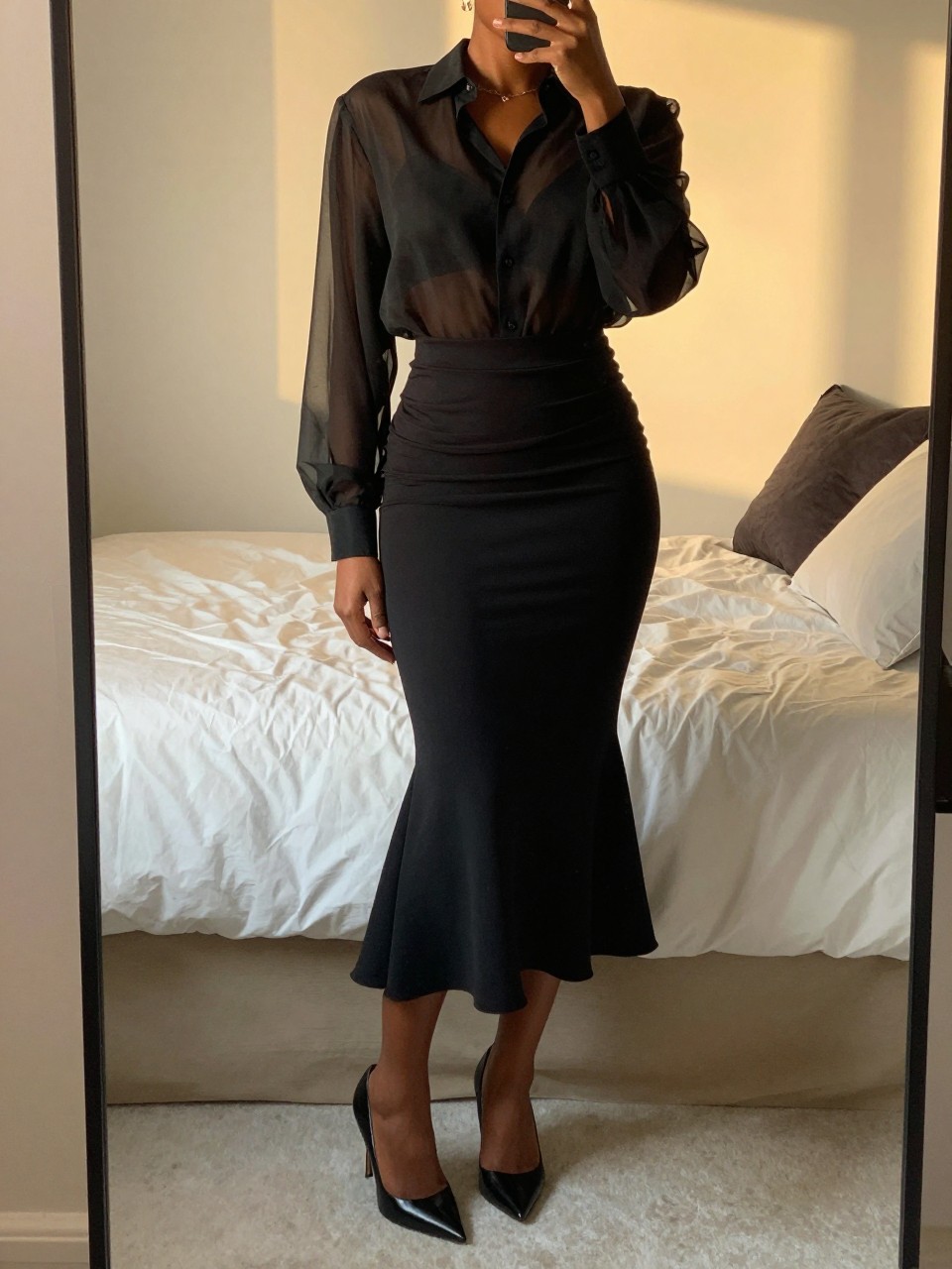mirror selfie of a black woman in a sheer black blouse and a black mermaid skirt with pointed pumps, standing with her weight on one leg, cropped at NECK so face doesn't show, in a feminine-themed bedroom, evening golden hour, iPhone photo quality.