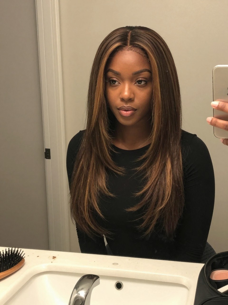Photo of a Black woman wearing a sleek dark lob with honey brown face-framing highlights, candid mirror selfie, at a bathroom vanity mirror, soft indoor lighting, containing a small lived-in detail such as a hairbrush on the vanity and a makeup bag nearby, latest iPhone photo quality.