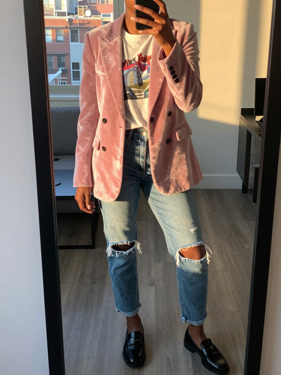 mirror selfie of a black woman in a pink velvet blazer over a graphic tee with ripped jeans and loafers, standing casually with phone near chin, cropped at NECK so face doesn't show, in a city apartment, evening golden hour, iPhone photo quality.