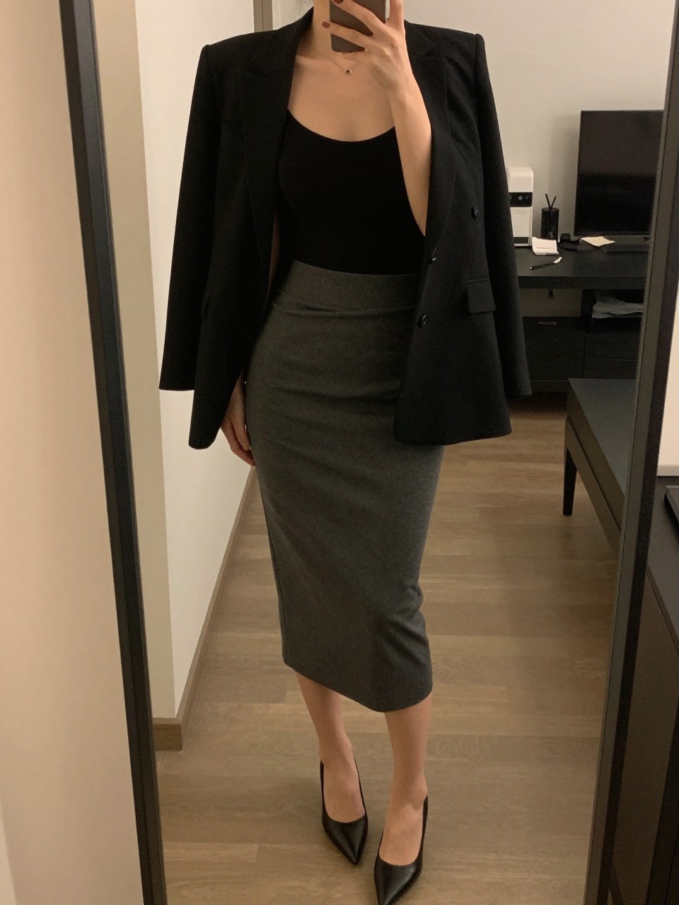 mirror selfie of a woman wearing a black bodysuit tucked into a charcoal gray midi skirt with a tailored black blazer and black pointed-toe pumps, one hand in her blazer pocket, cropped at NECK so face doesn't show, in a city apartment, warm moody lighting, iPhone photo quality.
