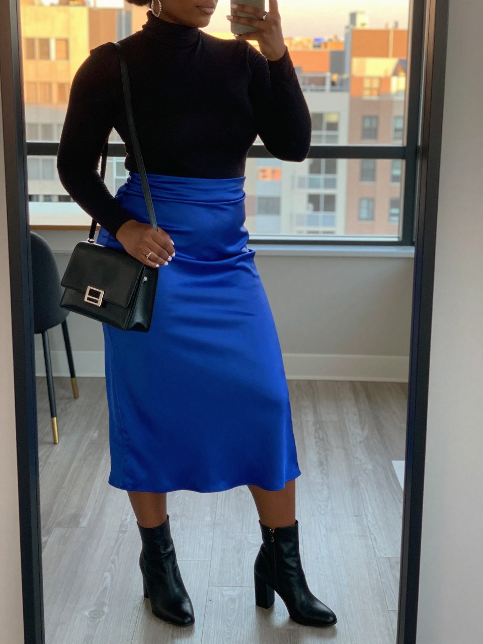 mirror selfie of a black curvy woman wearing a black turtleneck tucked into a royal blue satin midi skirt with black block-heel ankle boots and silver hoop earrings, holding a black crossbody bag with silver hardware, standing in an elegant pose, cropped at NECK so face doesn't show, in a city apartment, evening golden hour, iPhone photo quality.