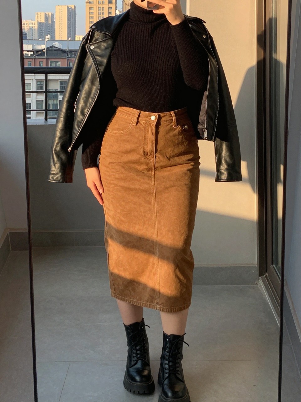 mirror selfie of a curvy woman wearing a black fine-knit turtleneck tucked into a caramel brown denim midi skirt with black chunky combat boots and a black leather jacket worn open, standing in an edgy pose, cropped at NECK so face doesn't show, in a city apartment, evening golden hour, iPhone photo quality.