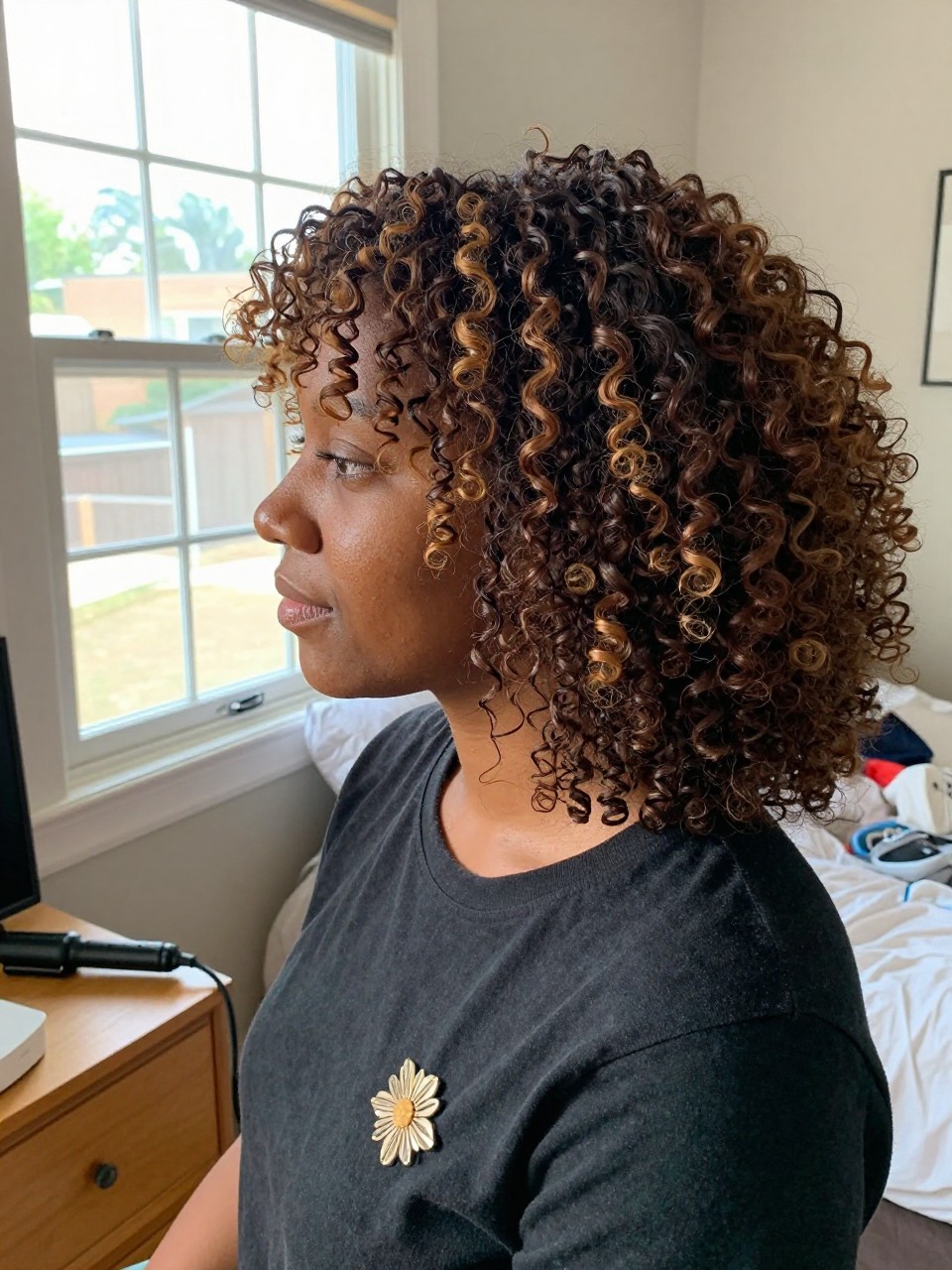 Photo of a Black woman wearing soft brown curls with mocha and toffee highlights, side profile view, in a sun-drenched bedroom, natural window light, containing a small lived-in detail such as a decorative pin on one side and a curling iron resting on the nightstand, latest iPhone photo quality.