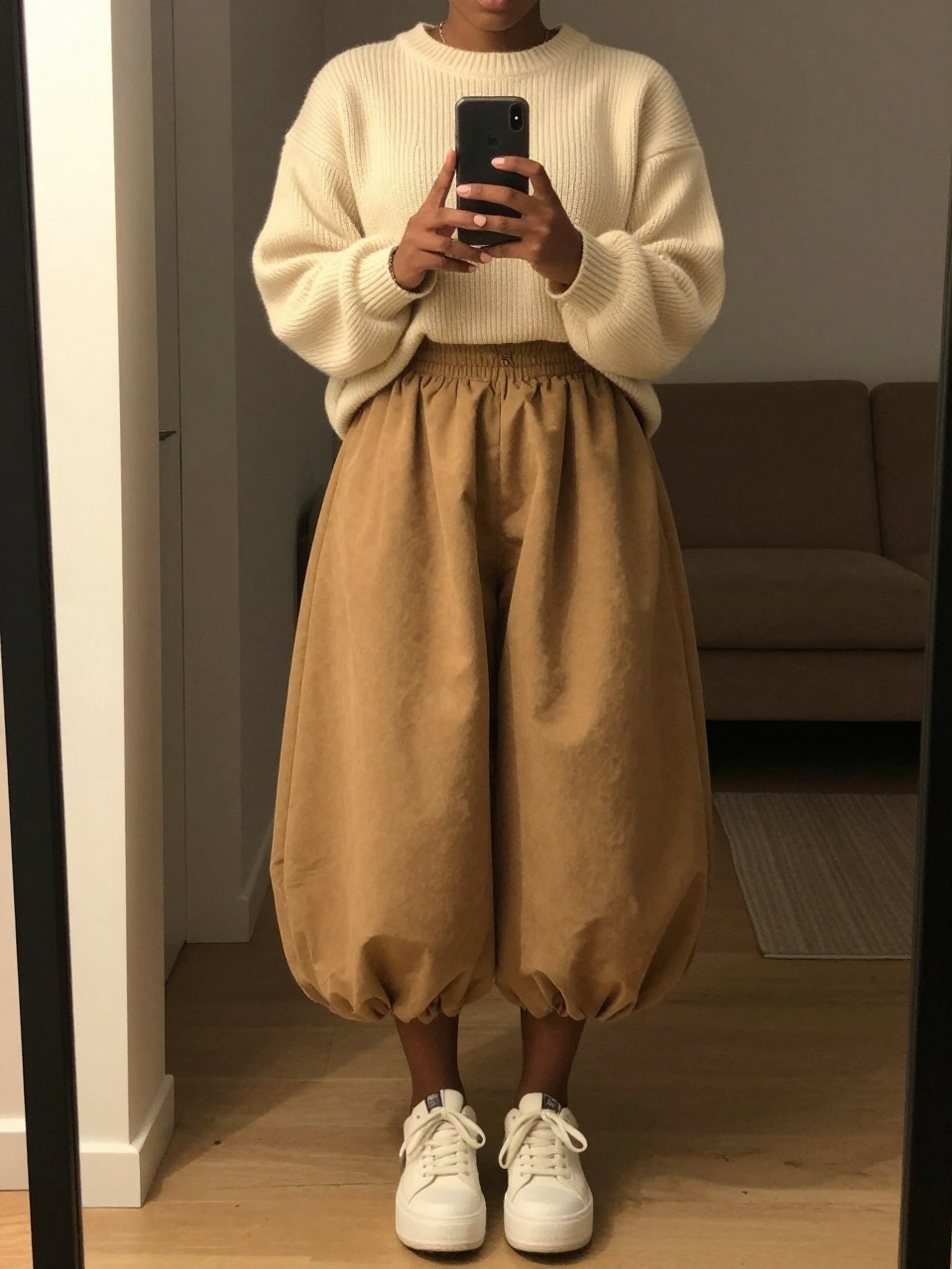 mirror selfie of a black curvy woman in an oversized cream sweater and a camel balloon skirt with platform sneakers, holding her phone at chest level, cropped at NECK so face doesn't show, in a city apartment, warm moody lighting, iPhone photo quality.