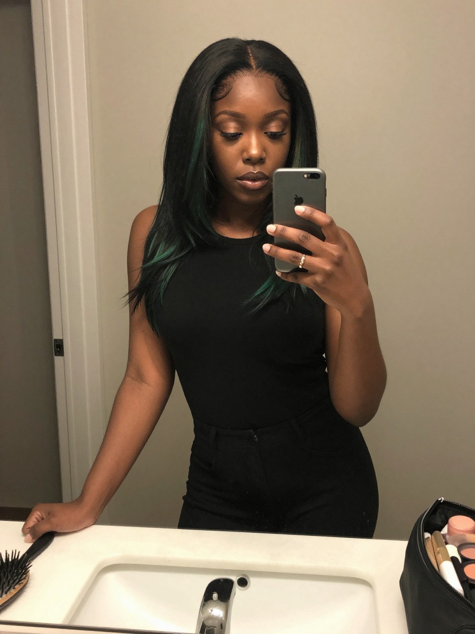 Photo of a black woman wearing a sleek black lob with green face-framing highlights, candid mirror selfie, at a bathroom vanity mirror, soft indoor lighting, containing a small lived-in detail such as a hairbrush on the vanity and a makeup bag nearby, latest iPhone photo quality.