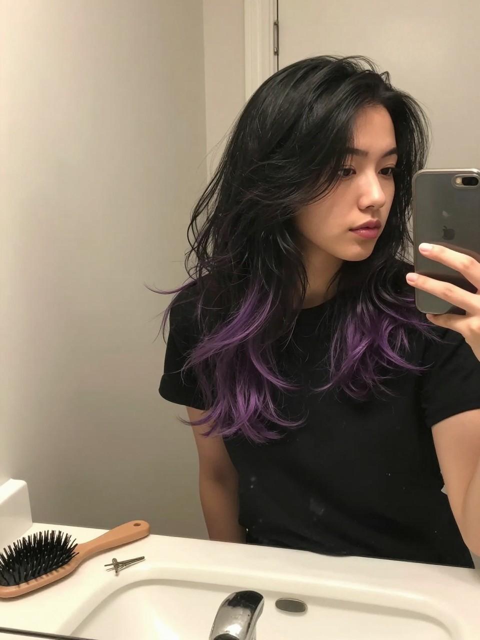 Photo of a woman wearing a wavy black lob with lilac purple highlights, candid mirror selfie, at a bathroom vanity mirror, soft indoor lighting, containing a small lived-in detail such as a hairbrush on the sink and a silver clip nearby, latest iPhone photo quality.