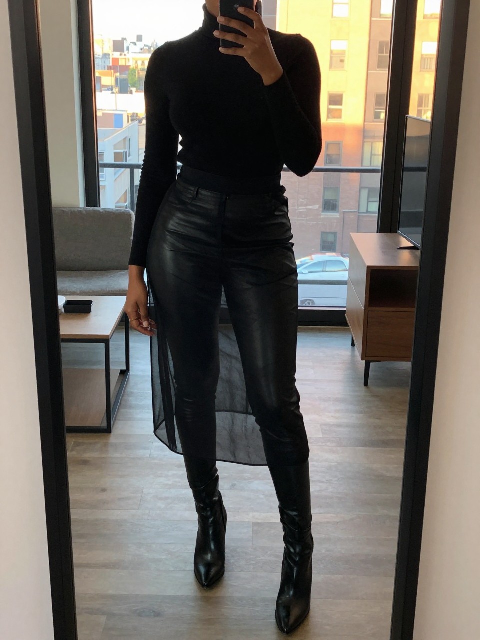 mirror selfie of a black woman wearing a black turtleneck with black leather leggings underneath a black sheer mesh skirt and black block-heel ankle boots, one hand adjusting her collar, cropped at NECK so face doesn't show, in a city apartment, evening golden hour, iPhone photo quality.