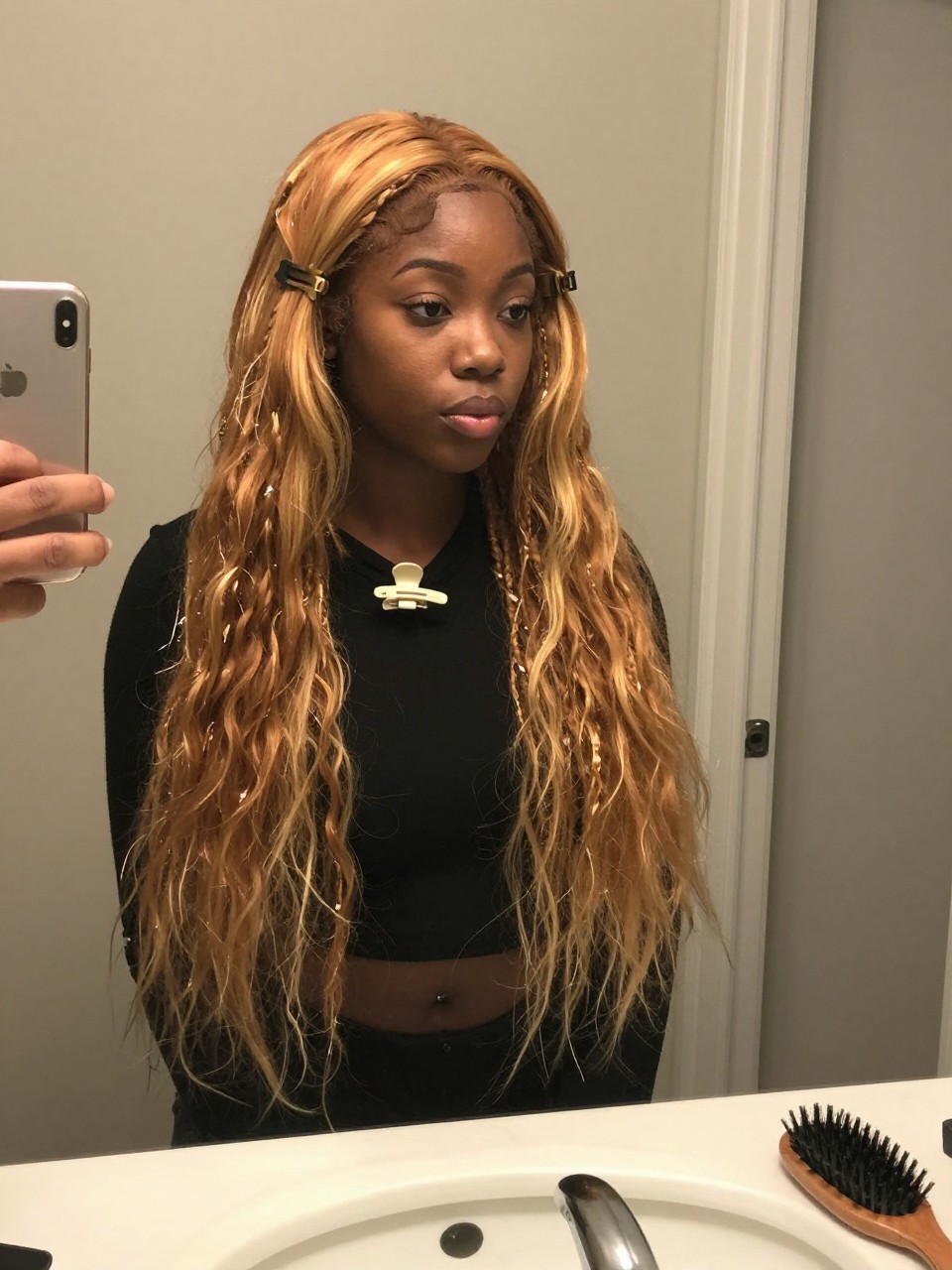 Photo of a black woman wearing a loose claw clip twist with ginger hair and warm blonde highlight pieces falling softly, candid mirror selfie, at a bathroom vanity mirror, soft indoor lighting, containing a small lived-in detail such as a claw clip clipped to the collar before use and a hairbrush on the sink, latest iPhone photo quality.
