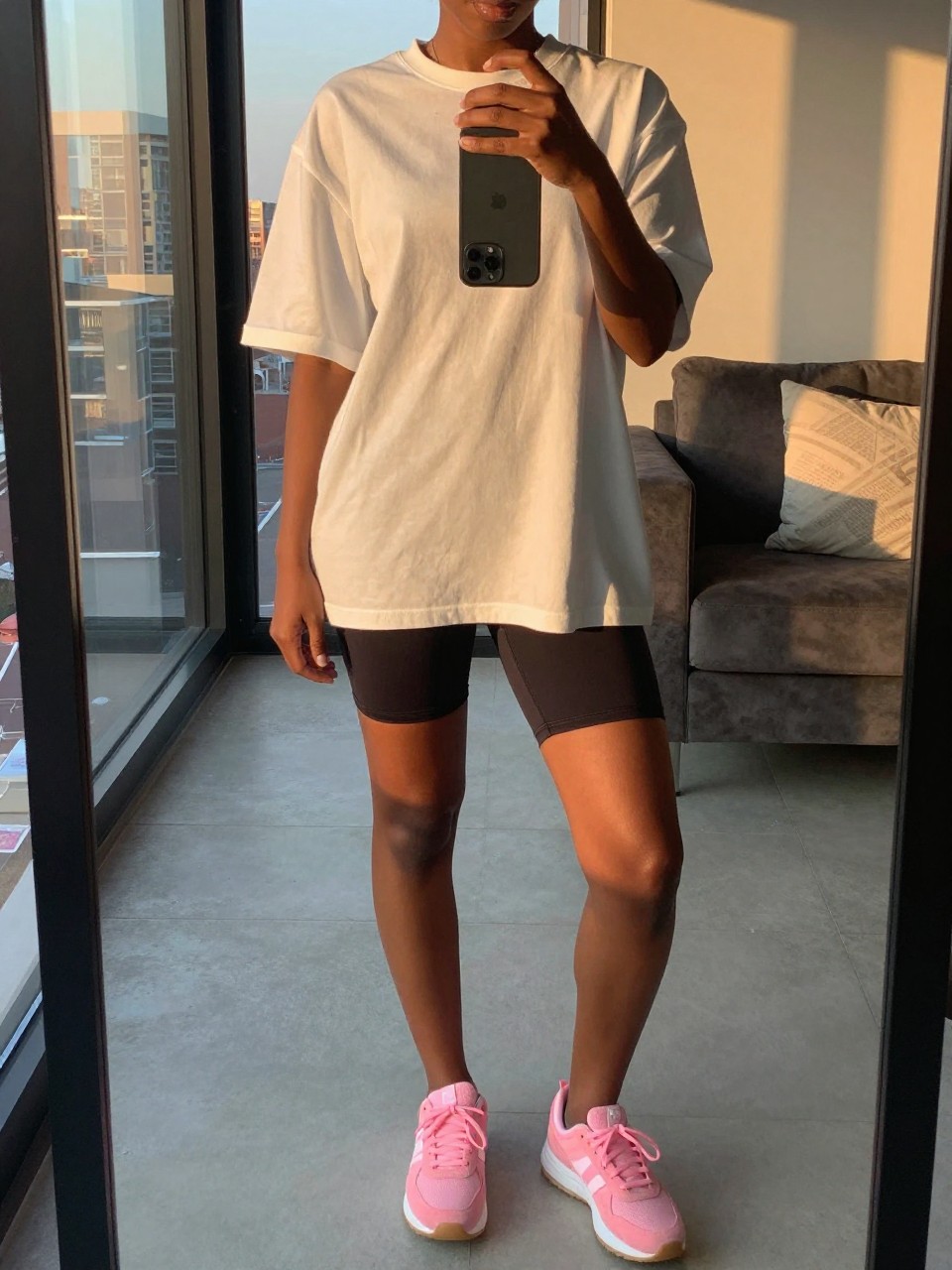 iPhone mirror selfie of a black woman in an oversized tee and bike shorts with pink sneakers, standing with phone near waist, cropped at NECK so face doesn't show, in a city apartment, evening golden hour, iPhone photo quality.