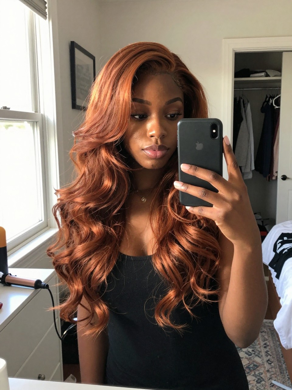 Photo of a black woman wearing mahogany copper balayage in soft loose waves, candid mirror selfie, sun-drenched bedroom setting, natural window light, with a curling wand on the dresser and a slightly open closet behind, latest iPhone photo quality.