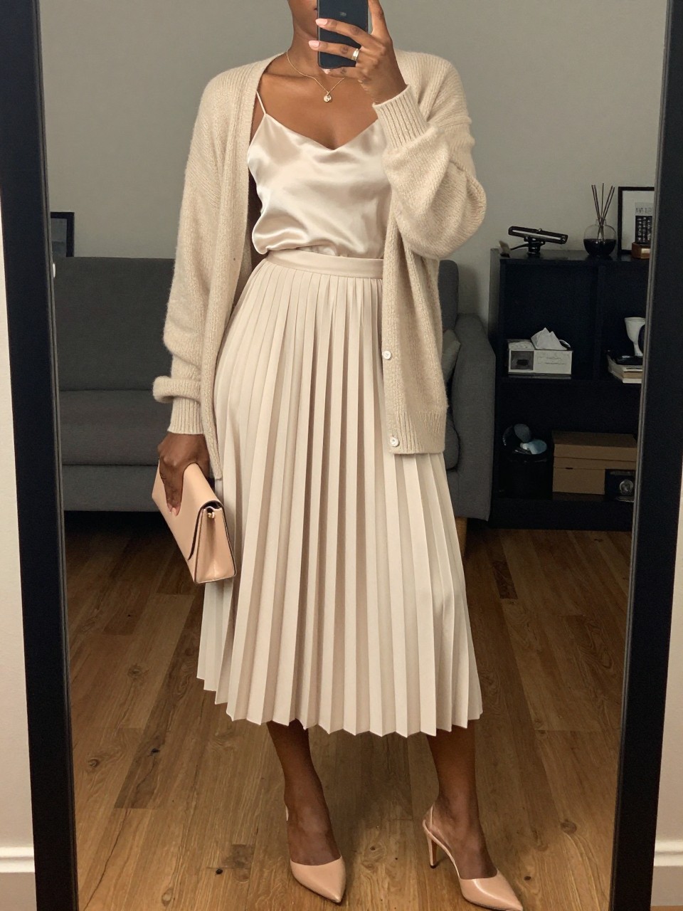 mirror selfie of a black woman wearing a champagne pleated midi skirt with an ivory satin cami and a soft beige light cardigan, nude heeled mules, and a blush small clutch, holding a phone in front of a mirror, cropped at NECK so face doesn't show, with a natural casual selfie pose, in a city apartment, warm moody lighting, including small lived-in details, latest iPhone photo quality.