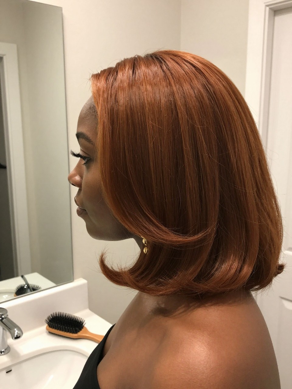 Photo of a black woman wearing soft copper brown hair in a smooth bouncy blowout, side profile view, bathroom vanity mirror setting, soft indoor lighting, with a round brush on the counter and statement earrings peeking through, latest iPhone photo quality.