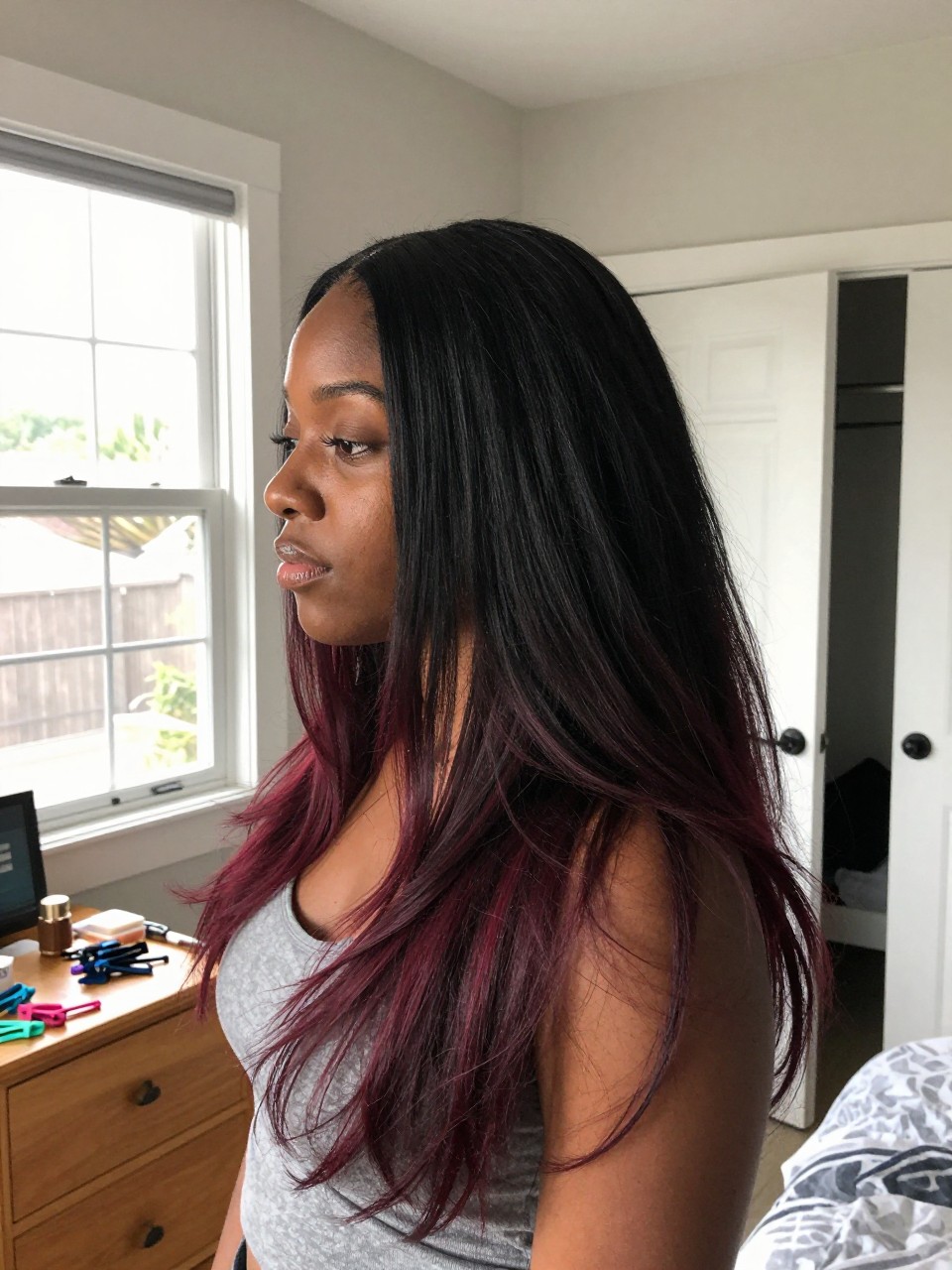 Photo of a black woman wearing long layered black hair with wine red highlights and soft movement, side profile view, in a sun-drenched bedroom, natural window light, containing a small lived-in detail such as sectioning clips on the dresser and a slightly open closet behind, latest iPhone photo quality.