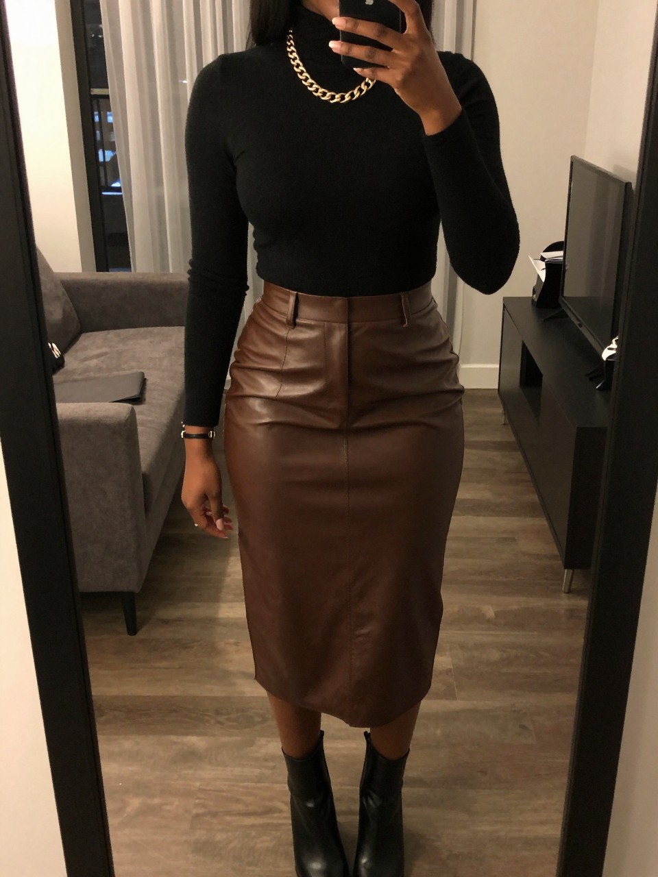 iPhone mirror selfie of a black woman wearing a black turtleneck tucked into a chocolate brown leather midi skirt with a chunky gold chain necklace and black ankle boots, one hand touching her necklace, cropped at NECK so face doesn't show, in a city apartment, warm moody lighting, iPhone photo quality.